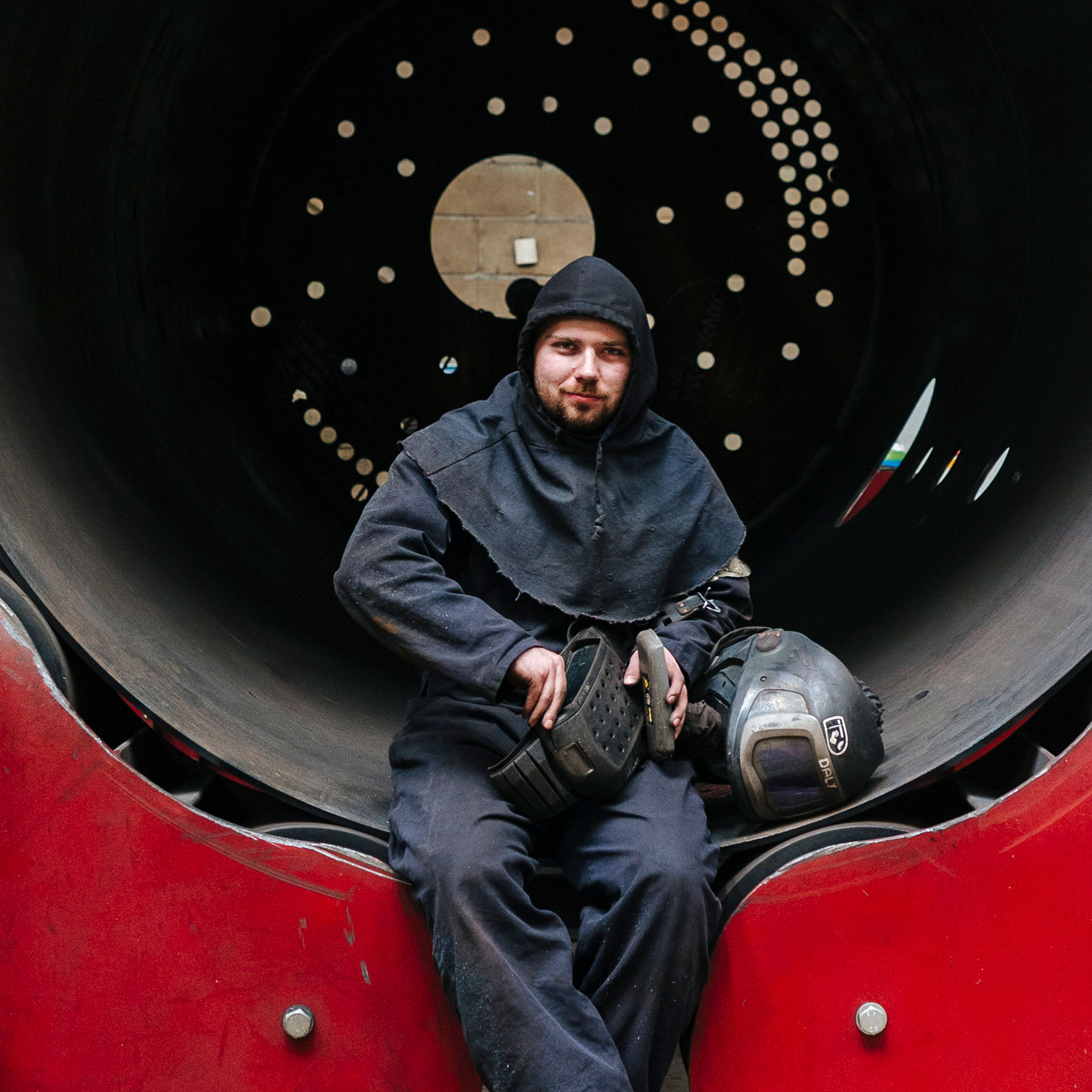 Man wearing overalls and safety wear, smiling, he is sat in a large boiler 